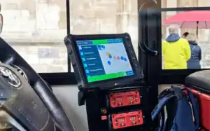 Vehicle-mounted touchscreen showing route and audio controls beside a bus driver’s seat