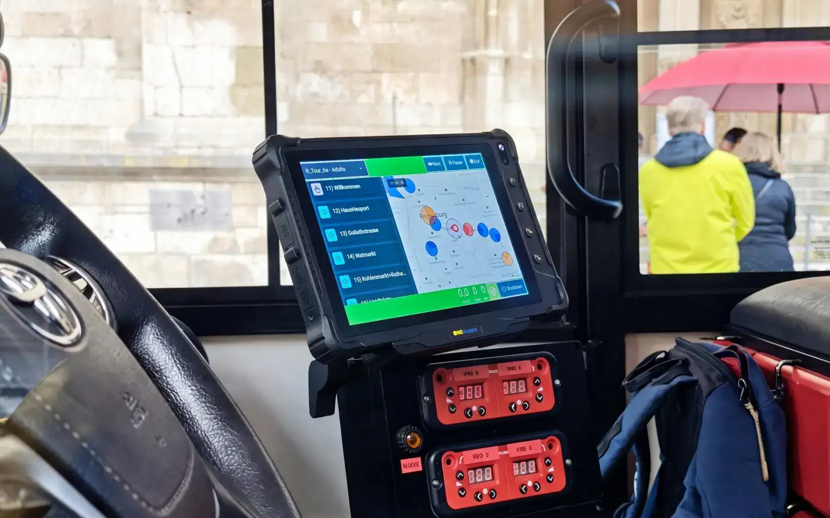 Vehicle-mounted touchscreen showing route and audio controls beside a bus driver’s seat